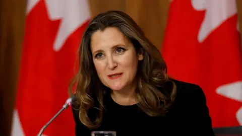 Reuters Chrystia Freeland, wearing a dark top, speaking into a microphone with a backdrop of Canadian flags.