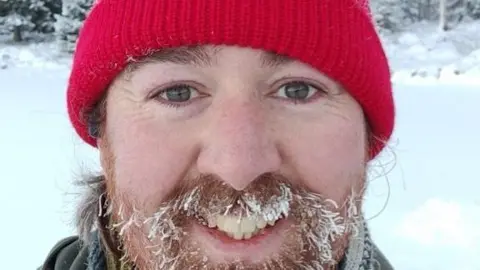 Oliver Eagle-Wilsher Oliver Eagle-Wilsher is smiling at the camera. He has a red beanie hat on. He is stood outside in the snow and has snowflakes on his ginger beard and hair. 