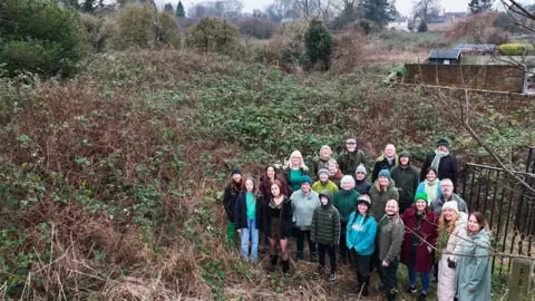 Tom Watson A photo taken at an angle looking down on the of the Friends of Driffield Sustainable Park - a campaign group of about two-dozen men and women standing on an overgrown patch of land in Driffield. They are dressed in winter clothing, surrounded by tall bushes and greenery, with garden fences to the right.