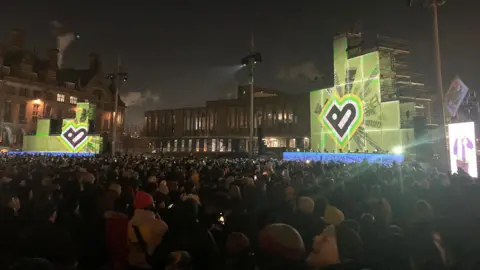 BBC Thousands of people wearing woolly hats standing in City Park in Bradford looking at two illuminated stages lit up with a big letter B in black surrounded by a yellow, green and pink background.