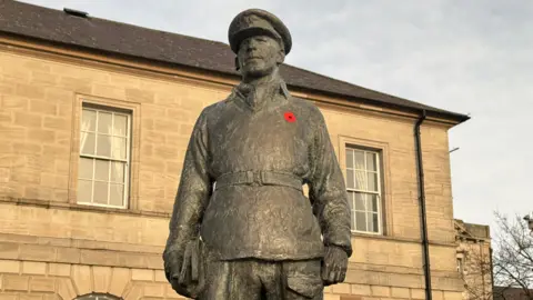 A statue honouring Mayne - it is a bronze figure with its arms at its side wearing a military uniform including a peaked cap. A red poppy is on its left breast. In the background is a sandstone building.