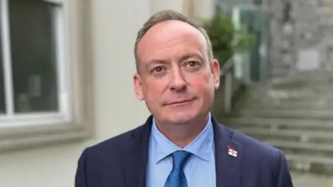 BBC Politician in blue shirt and blazer smiling at camera