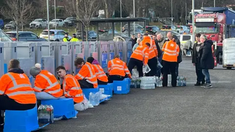 BBC Picture of staff members in orange hi-viz jackets sat on blue crates and surrounded by bottles of water at the water station in Zip World Conwy, with queue of cars in background and