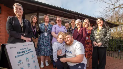 City of Wolverhampton Council A group of 11 adults and one child standing outside a red-brick building on a red-tiled floor. The man, wearing tracksuit bottoms and T-shirt, is crouched with his young daughter in his arms. The other adults, all female, stand around him. A park can be seen to the right behind them.