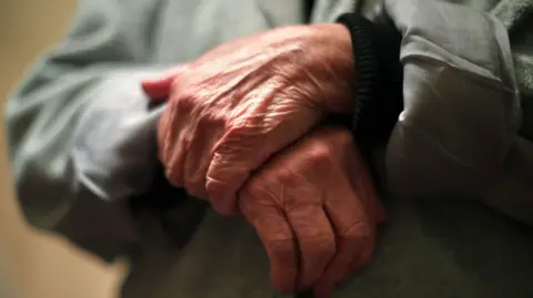 PA Media A close-up image of the hands of an elderly person, they are crossed over in front of their body. Their arms are covered with the sleeves of a grey jacket, which is slightly out of focus. A beige-coloured wall is in the background.