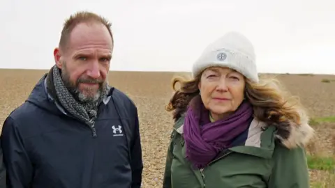 Ralph Fiennes and fellow campaigner Fiona Gilmore pictured on the beach at Aldeburgh in Suffolk. He has short, light brown hair and a greying beard, wearing a black waterproof jacket and black and white scarf. Fiona has long, wavy, auburn hair, wearing a white woolly hat, purple scarf and khaki green waterproof coat, with a fur-lined hood. The sky is cloudy and they are standing on shingle.