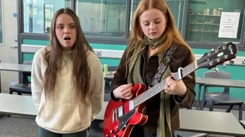 Ebony (left) is singing and Olivia (right) is playing a red guitar. Ebony is wearing a white hoody and green leggings and Olivia is wearing a brown jacket and green scarf. There are tables and chairs in the classroom behind them.