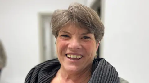 Anna Forgaard has short hair and is wearing a scarf. She is smiling and showing her teeth. She is in focus and the background is blurred.