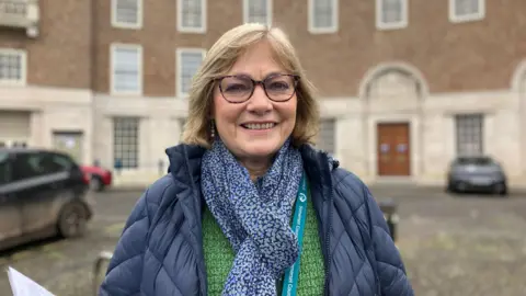 BBC Sarah Wakefield  is standing in front of the council building. She is wearing a blue scarf and a blue jacket. She is smiling at the camera.