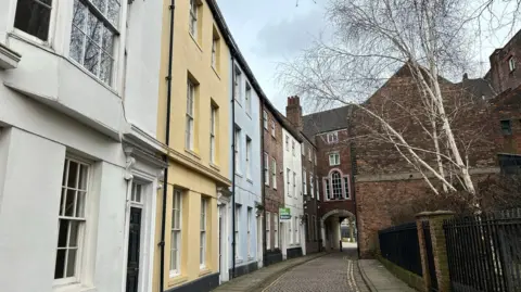 Charlie O'Loughlin/ BBC A narrow, cobbled street lined with historic townhouses painted white, yellow and pale blue. The buildings have large windows. A brick archway connects two buildings in the distance. A leafless birch tree stands behind railings on the right and the sky is blue with light clouds.
