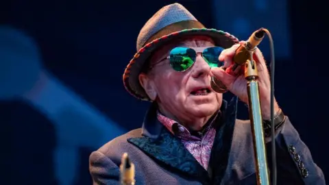 Getty Images Van Morrison on stage. He is wearing a hat, a jacket and a patterned pink shirt. He is also wearing sunglasses and holding a microphone.