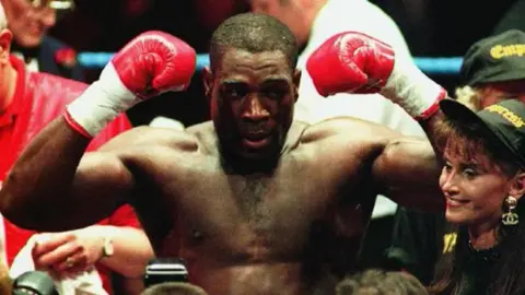 PA Media Frank Bruno stands in a ring in the 1990s with his arms raised in triumph, wearing red boxing gloves.