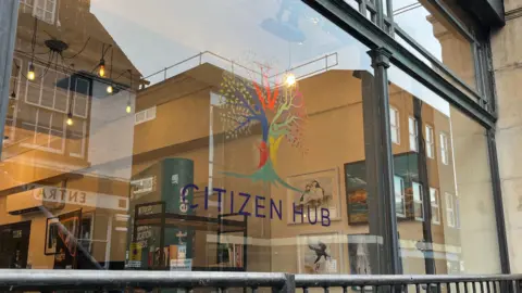 Tom Jackson/BBC A window looking into the Citizen Hub. There is a logo and the words Citizen Hub printed on the glass. Inside there are photos of the wall, paintings and bookshelves.