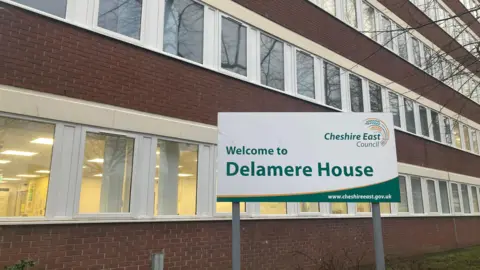 An angled shot of Delamere House in Crewe, Cheshire East Council's headquarters, with it's brick facade interlaced with long sections of white windows. The Delamere House green and white sign is pictured on the lawn in front of the building.