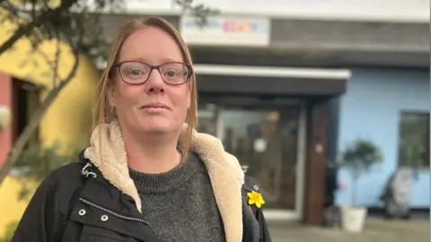 Fiona Irving/BBC Kerry is wearing a grey jumper, black coat and has a bag over her right shoulder. The centre in the background is painted yellow and red on the left, and blue and yellow on the right.