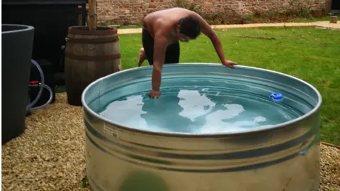 Sivo Wellness A man wearing shorts carefully tests the temperature of an ice bath. It is a large stainless steel bath, very nearly full of water. It stands on a patch of gravel in front of a grassy lawn.