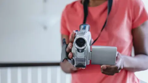 Getty Images Stock shot of torso of anonymous black young person holding a video camera pointing directly to camera