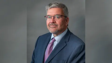 Cheshire East Council A headshot of Nick Mannion, the Labour leader of Cheshire East Council. He has short grey hair and is wearing clear glasses with a reddish brown frame, a dark blue suit, light blue shirt and purple patterned tie.