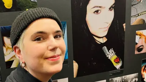 AJ Turner AJ Turner wearing a black beanie and has short blonde hair taking a picture with a an image of her younger self at the exhibition.