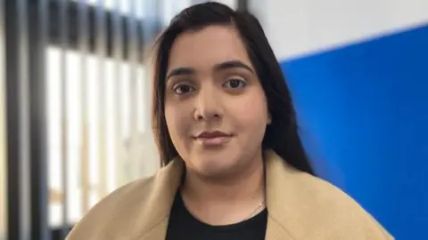 BBC/Michelle Lyons Marium Zameer, wearing a beige coat and black top, standing in front of a blue wall and, to her left, a window with blinds open.