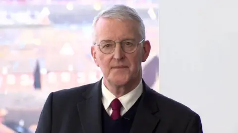 BBC A grey haired man wears circular glasses, as well as a dark suit jacket, a dark jumper, a white collared shirt and a red tie. He is speaking from a podium, and behind him are blurred rooftops of nearby buildings.