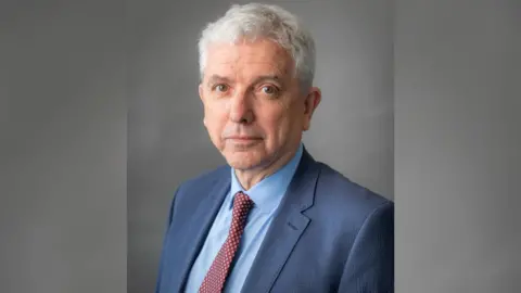 Cheshire East Council A headshot of Michael Gorman, the deputy leader of Cheshire East Council and the leader of the Independent Group. He has white hair and is wearing a dark blue suit, light blue shirt and red tie with cream spots.