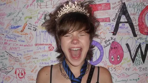 LottiePaterson/MuseumOfYouthCulture A girl with a pixie cut and brown hair wearing a tiara. She appears to be screaming with her eyes closed and also wears a necktie and silver necklace.
The background is full of colourful scribbles and messages.