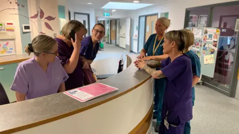 Mark Ansell / BBC Three women in scrubs behind a nurses station desk and three women in scrubs leaning on the desk on the other side