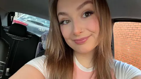 Layla Cahill Harris Layla Cahill Harris smiles into the camera. She is wearing a pink and white t-shirt and is in the car. She has long brown hair which frames her face, pink lipgloss on and a nosering.