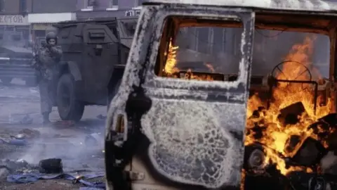 Getty Images A burning van on a city street during the Troubles. Part of the back of the van is open and flames are consuming the inside, the outside of the van is scorched by the heat. In front of the van are two Army vehicles and a soldier with a rifle is standing beside it with a had towards his mouth to block out the smoke.
