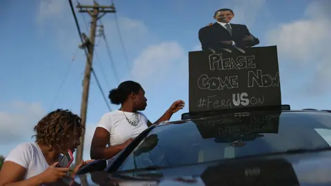 Getty Images Demonstrators carry a sign with a picture of Barack Obama reading, "Please Come Now," on August 17, 2014 in Ferguson
