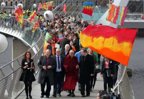 AFP The Dalai Lama and a large crowd of people cross the Peace Bridge which is decked with colourful flags