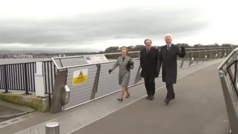 Pat and John Hume and Bill Clinton walk across the Peace Bridge