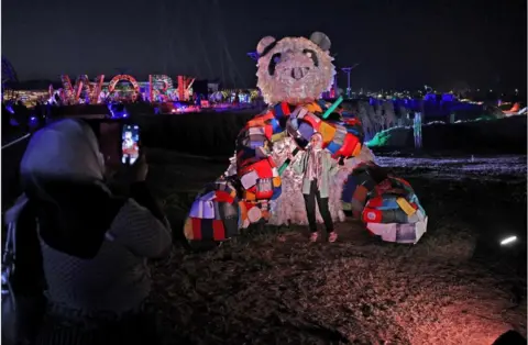 AFP A woman is taking a photo in front of a big multicoloured panda. It is night time and there are lights in the background.