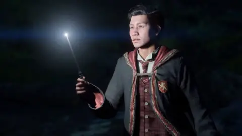 WB GAMES A student of East Asian appearance wears the black robes of a hogwarts student, emblazoned with a red shield with a golden lion on it denoting membership of Gryffindor house. A thread holds the robe together at chest level, and we can see the garment's velvety red lining and golden trim. In one hand he holds a wand between his thumb and forefinger, and the tip is brightly illuminated. The wand provides the only source of light in the otherwise pitch black scene, and it casts a spooky white glow on the student's body.
