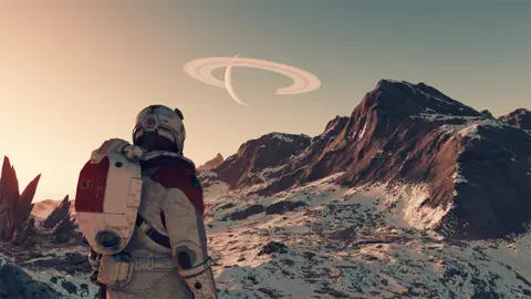 Bethesda A screenshot from Starfield shows the back of an astronaut character in spacesuit and helmet surveying a barren planetscape. It is rocky, with cliffs jutting out of the ground. The rocks are covered with a white powder - either dust or snow. In the distance another planet, and its horizontal rings, can be see in the sky.