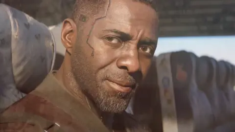 CD PROJEKT RED A computer-generated Idris Elba in close-up, sitting in the window seat of a train carriage. His short, stubbly beard is illuminated by sunlight from outside and he has a serious, thoughtful expression. Some sort of wire/track runs from his temple to his cheek, giving him a futuristic appearance.
