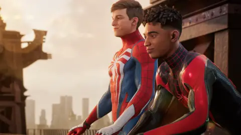 Marvel A screenshot from Spider-Man 2 showing protagonists Peter Parker and Miles Morales sitting on a balcony. They're both wearing their figure-hugging Spidey Suit costumes, but neither is wearing a mask. Peter's suit is mostly red, with thin black lines suggesting a spider's web. It also has blue panels on the biceps, triceps and flanks of his torso. A large, white, spider decal is printed on his chest, its legs extending to the edges of the suit. Peter is white, with fairly short, neatly cropped hair. Mile's suit has a similar design but is mainly black, with red panelling, web lines and spider decal. He's a young black man with short, afro-style hair style in locs. Both look out over New York contemplatively.