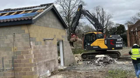 Kate Bradbrook/BBC A digger is demolishing the Captain Tom spa building