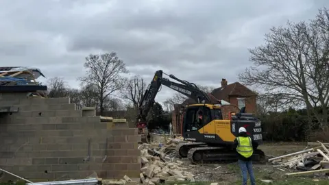 Kate Bradbrook/BBC A digger is demolishing the Captain Tom spa building