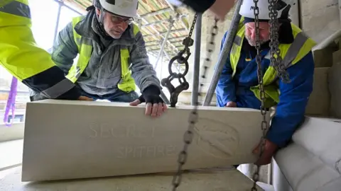 Finnbarr Webster Men wearing high-vis and hard hats putting a stone with a spitfire carving in place