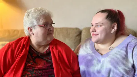 BBC A woman in a bright red cardigan and a younger woman in a purple top sitting on a sofa together smiling at each other