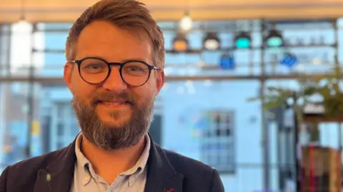 Harriet Heywood/BBC Alex Hughes is standing inside the community hub. He has a beard and circle-framed glasses and is wearing a navy blazer and a light blue shirt. He is smiling at the camera