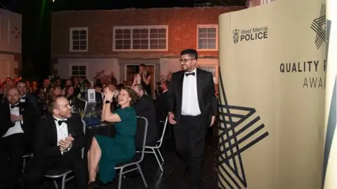 West Mercia Police Men and women in black tie and evening dresses are sitting at tables, applauding as the awards are announced at a ceremony. A big poster with "West Mercia Police" on it sits to one side.