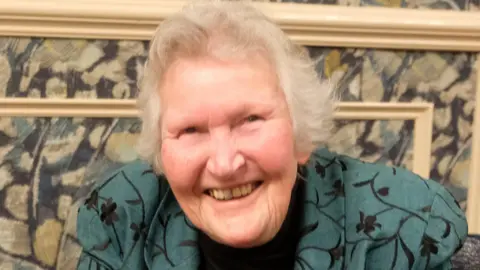 Paul Buchanan Gill Green, smiling to the camera, sitting down, wearing a black top, cat broach and a green and black jacket. She has short grey hair.
