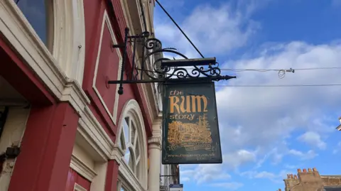 Sign saying "The Rum Story" attached to red brick building. The sign is green, the lettering orange and the image is often several ships.
