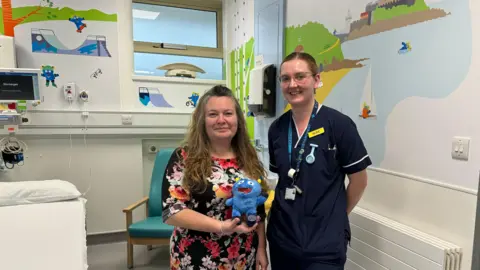 Courtney Sargent/BBC Clare Backlund-Leale is wearing a red and black flowery dress and is holding a small blue teddy bear. Alice Wary is wearing dark blue scrubs and they are both standing in front of a mural. Will can be seen in the mural behind them.