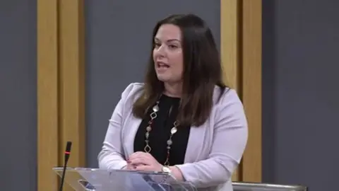 Vikki Howells in the Senedd. She is stood at a lecturn about to speak. She is wearing a black top underneath a lilac jacket and has a beaded and gold necklace on. Her dark shoulder-length hair is untied.