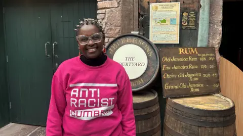 BBC Janett Walker in pink jumper which says Anti Racist Cumbria. She has large colourful glasses and is smiling at the camera. She is standing by barrels and a wooden sign listing different types of Jamaican rum.