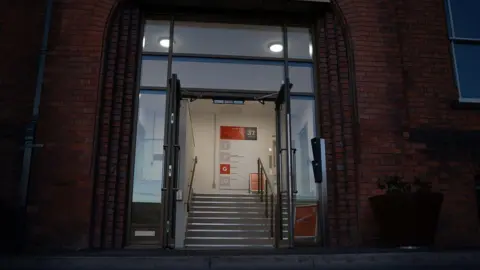 Wyre Forest District Council A dark shot of the entrance to a building. It is a former court and has red brick. But there are modern doors. There are stairs and signs on the wall telling visitors where to go.